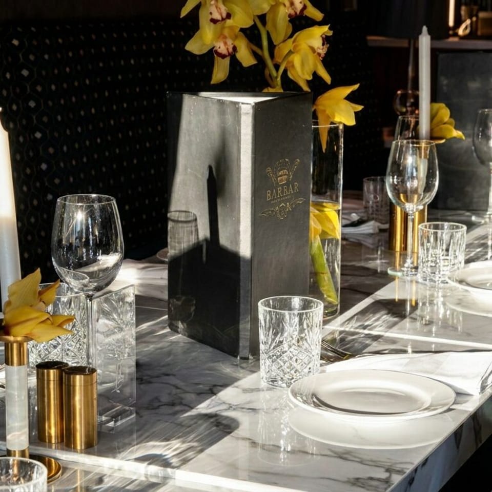 Elegant restaurant table set with white plates, crystal glassware, candles, yellow flowers, and a Barrar menu on marble.