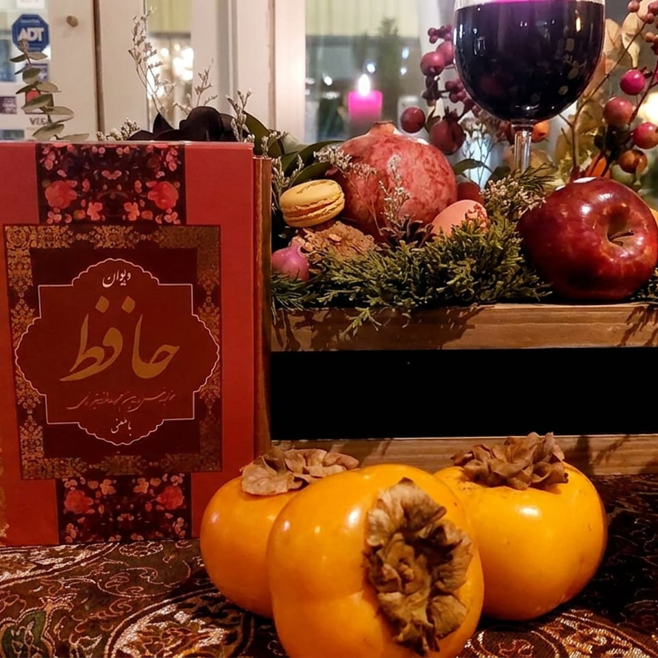 Persian poetry book surrounded by pomegranates, persimmons, apples, flowers, and a decorative wineglass on a patterned table.