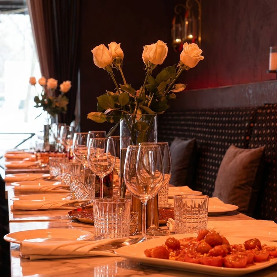 Elegant restaurant table set with wine glasses, orange roses, candles, and a plated pasta dish beside a cushioned banquette.